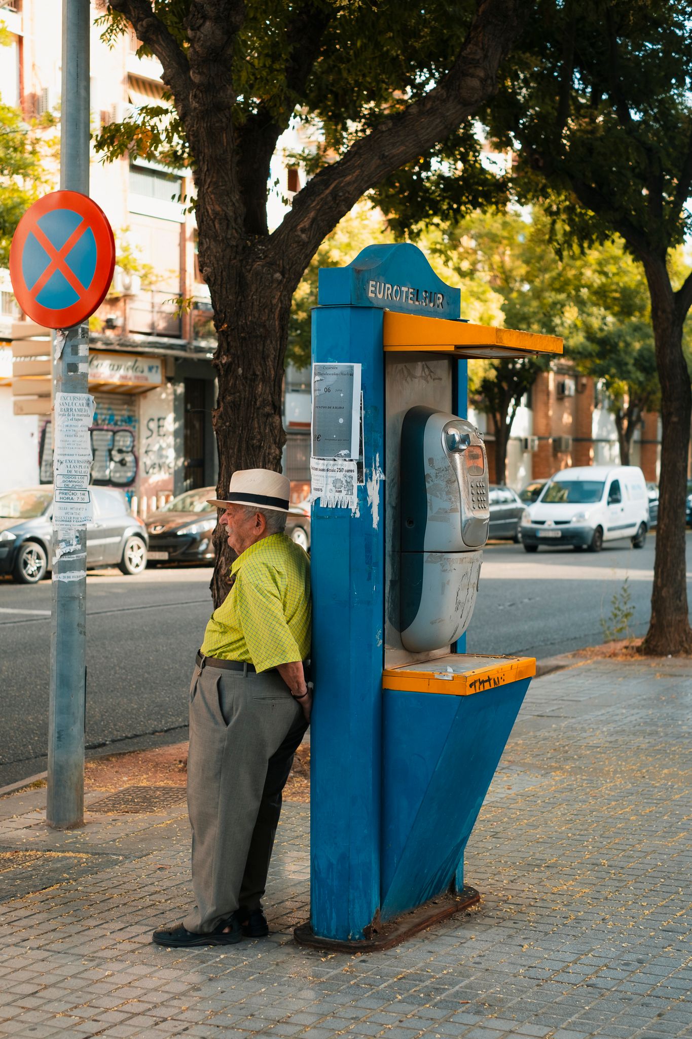 An elderly man in a yellow shirt resting against a blue payphone booth on a tree-lined street