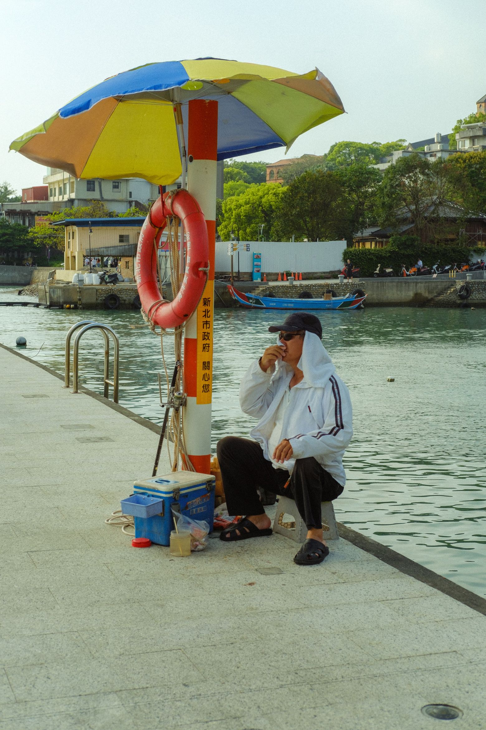 A vendor crouched under a colorful striped parasol beside a harbor railing at the waterfront