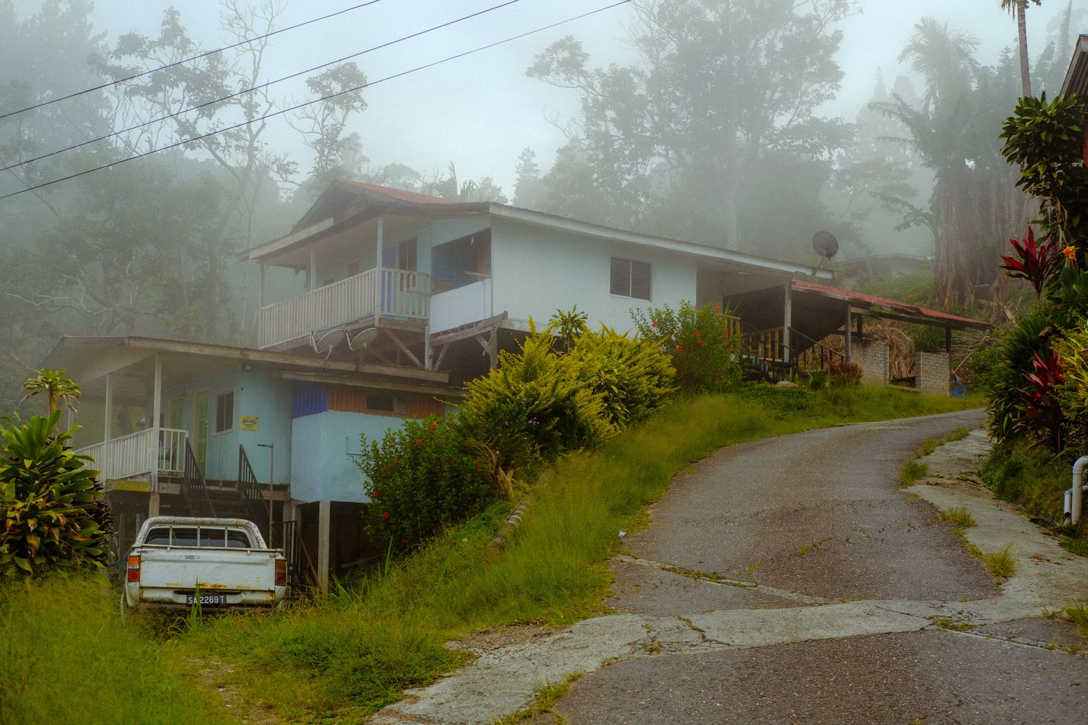 A car parked on a misty hillside lane beside houses in a foggy mountain village