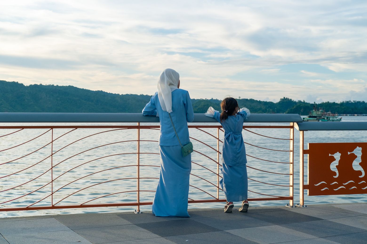 Two women in long dresses leaning on a railing, looking out over a lake and hills