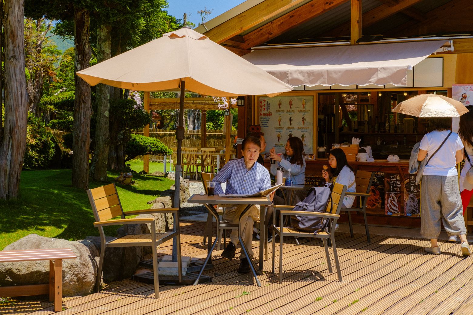 People relaxing at a wooden café terrace shaded by a white parasol among trees