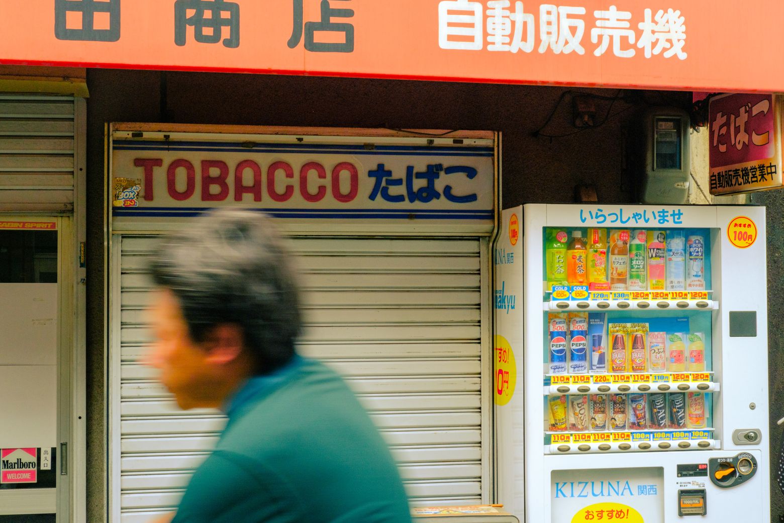 A man walking past a tobacco shop and a row of brightly lit Japanese vending machines