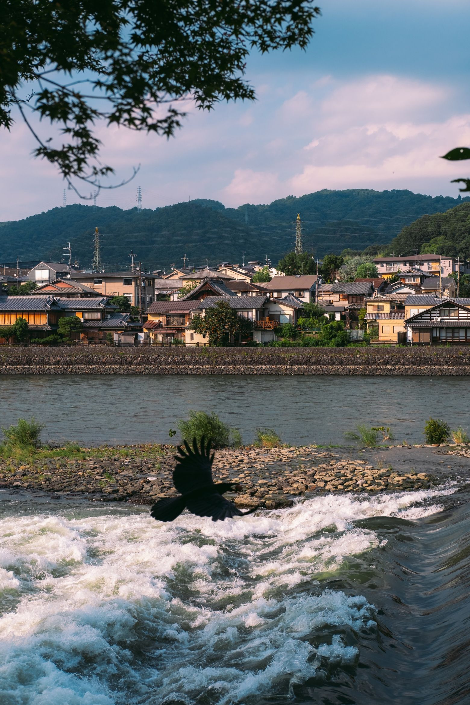 A riverside town below green mountains, with water rushing over a weir in the foreground