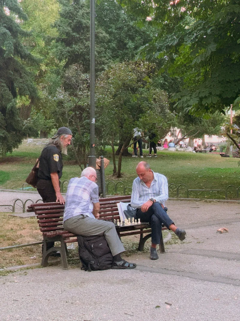 Two men playing chess on a park bench while a third watches