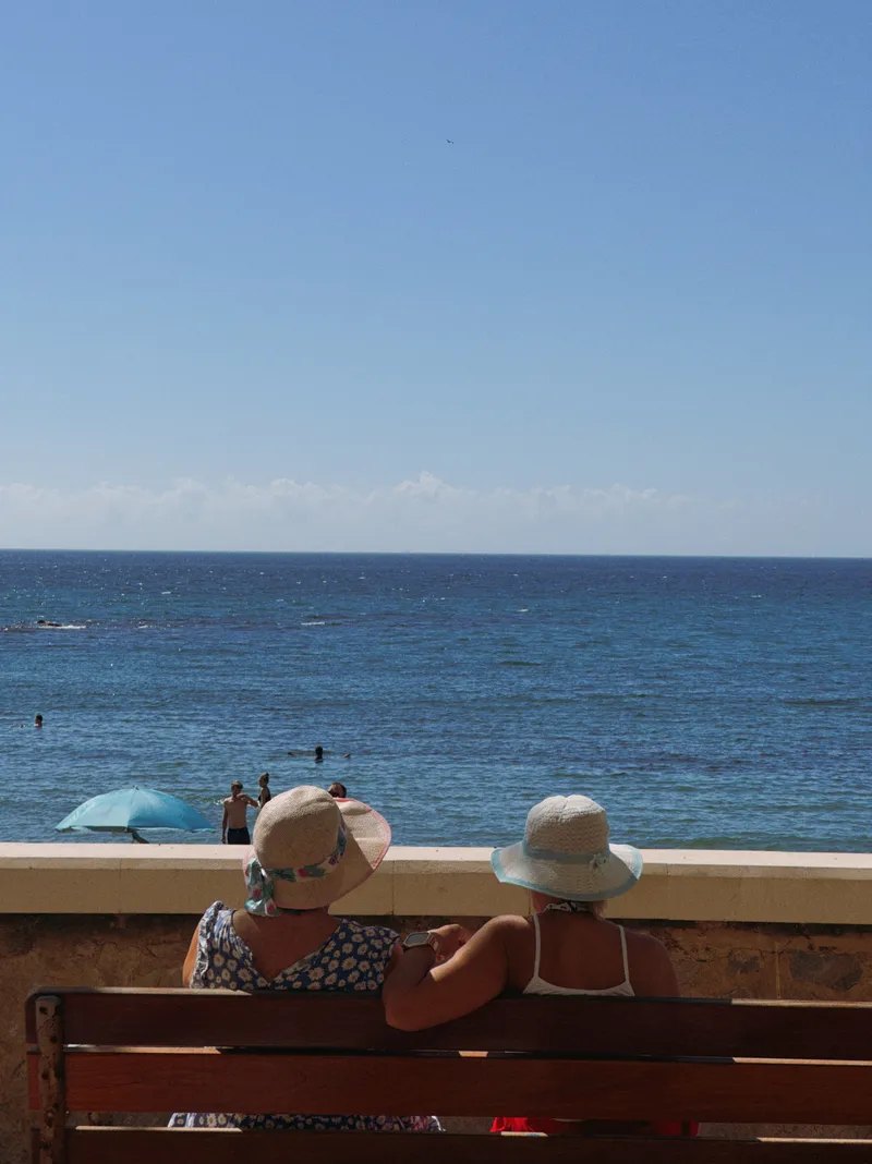 Two women in sun hats sitting on a bench, looking out at the sea